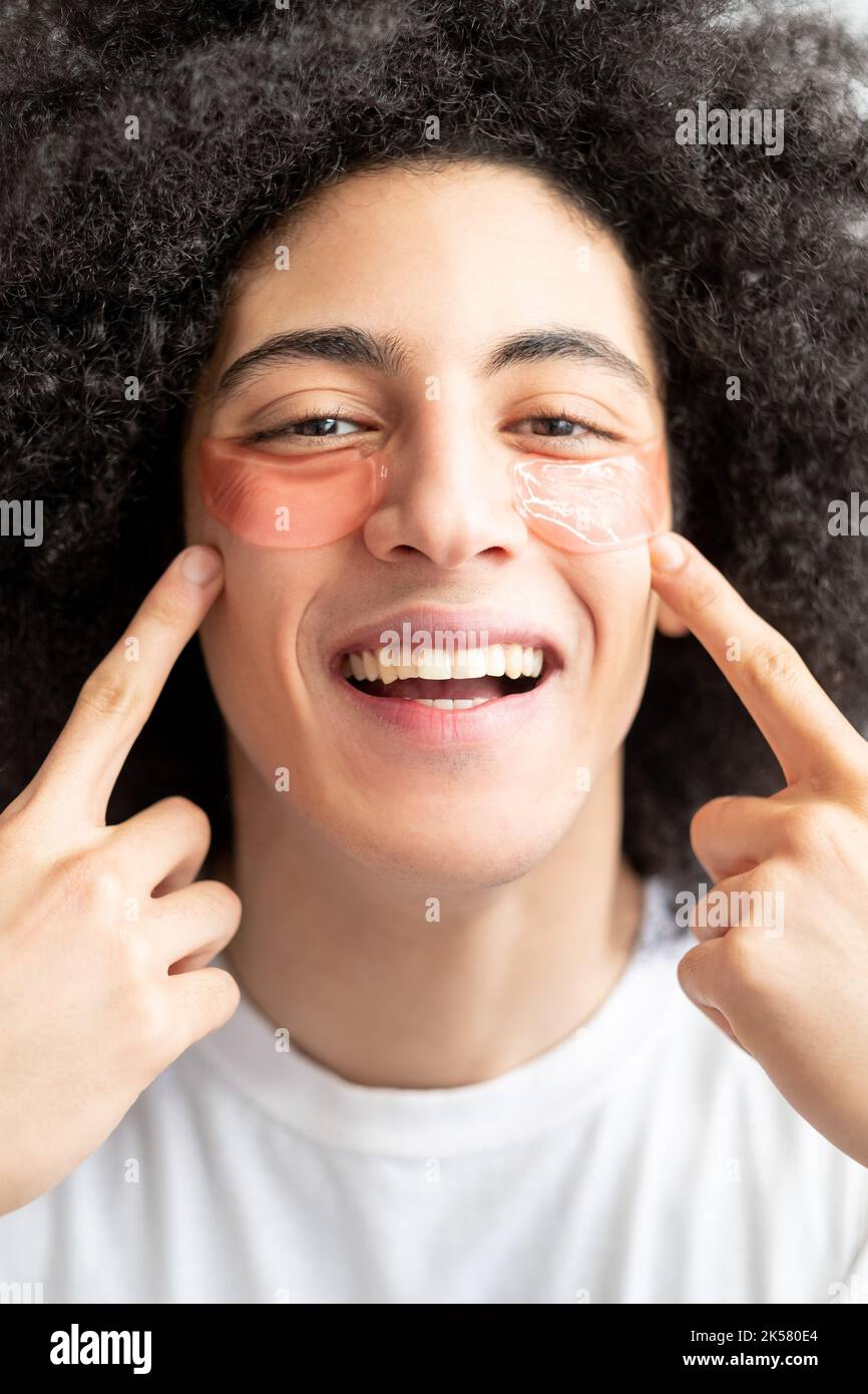 Soin de la peau pour homme. Produit cosmétique. Toilettage amusant pour hommes. Joyeux gars avec un sourire toothy brun curly cheveux pointant vers des taches roses d'oeil d'hydrogel sur la perf Banque D'Images Soin de la peau pour homme. Produit cosmétique. Toilettage amusant pour hommes. Joyeux gars avec un sourire toothy brun curly cheveux pointant vers des taches roses d'oeil d'hydrogel sur la perf Banque D'Images