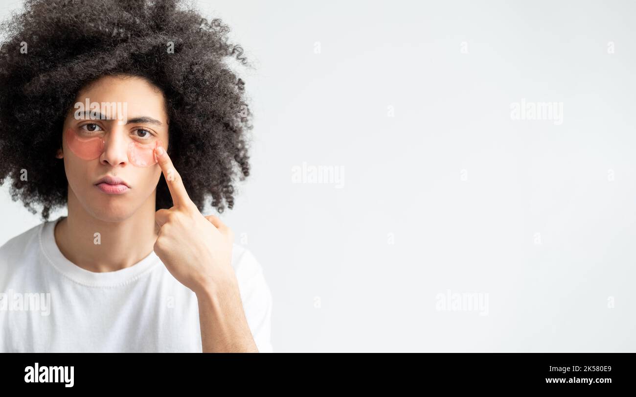 Soins du visage pour hommes. Procédure anti-âge. Mode de vie métrosexuel. Mécontent mec fatigué avec des cheveux bruns bouclés pointant vers des timbres roses pour les yeux hydrogel Banque D'Images Soins du visage pour hommes. Procédure anti-âge. Mode de vie métrosexuel. Mécontent mec fatigué avec des cheveux bruns bouclés pointant vers des timbres roses pour les yeux hydrogel Banque D'Images