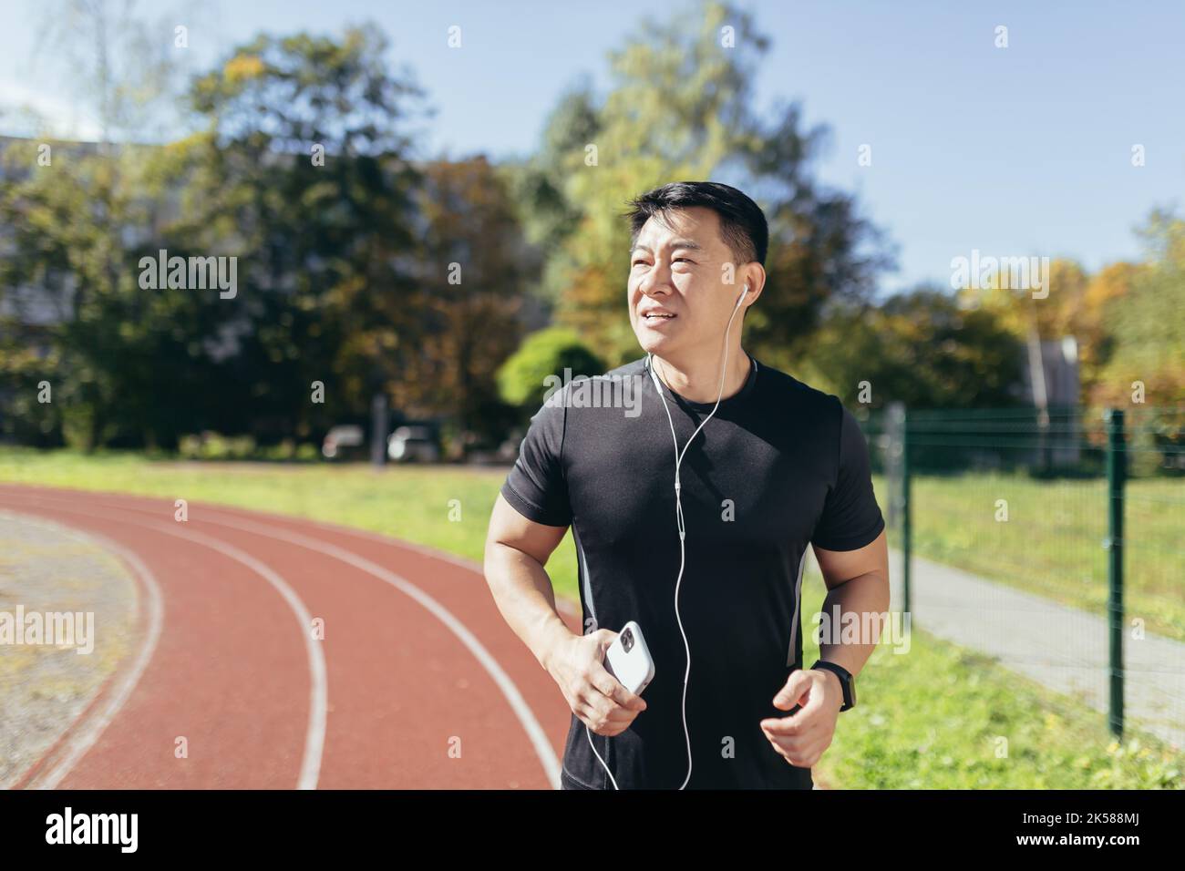 Un sportif asiatique heureux et réussi qui court le matin, un jour ensoleillé au stade, un homme avec un casque filaire qui écoute de la musique avec un smartphone et des podcasts avec des livres audio. Banque D'Images
