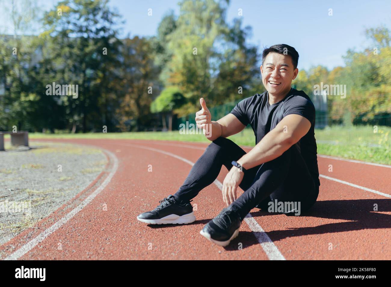 Portrait d'un sportif asiatique assis sur un tapis roulant de stade dans un parc public par une journée ensoleillée d'été, l'homme regarde l'appareil photo et sourit en donnant des pouces, coureur de vêtements de sport à l'extérieur. Banque D'Images