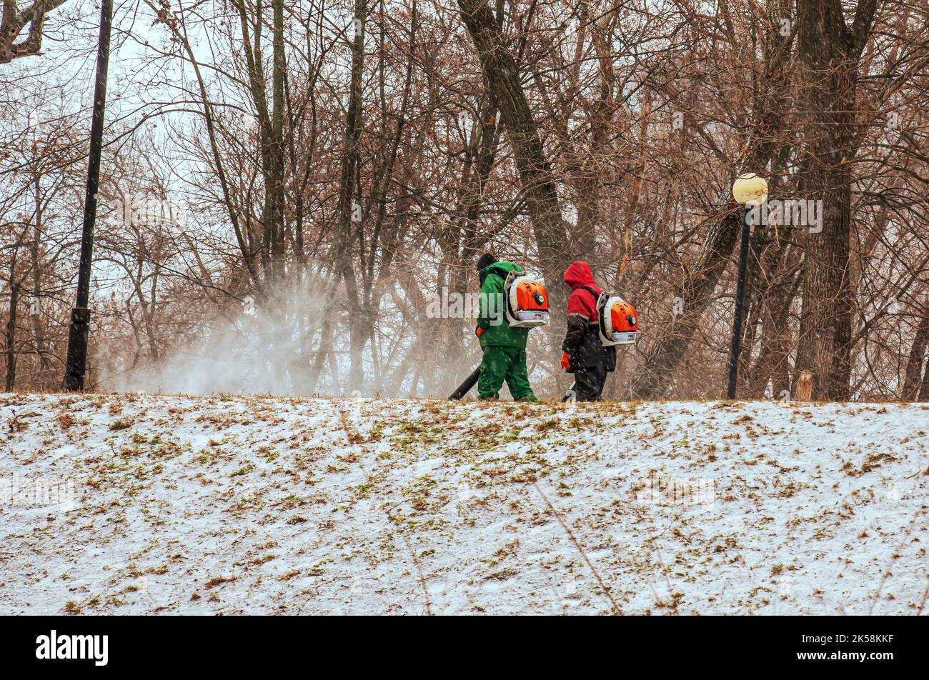 Dnepropetrovsk, Ukraine - 02.04.2022: Travail saisonnier des services publics de la ville dans le parc. Un travailleur équipé d'un souffleur de sac à dos motorisé souffle la neige d'un ap de stationnement Banque D'Images