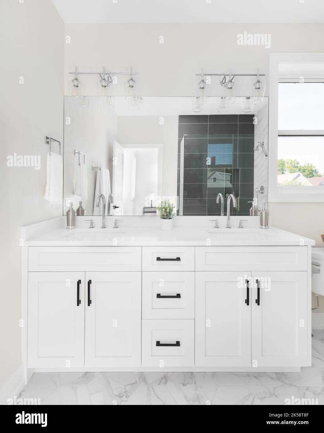 Une salle de bains avec meuble-lavabo blanc, comptoir en marbre blanc et robinets argentés avec vue sur la douche carrelée à travers le miroir. Banque D'Images