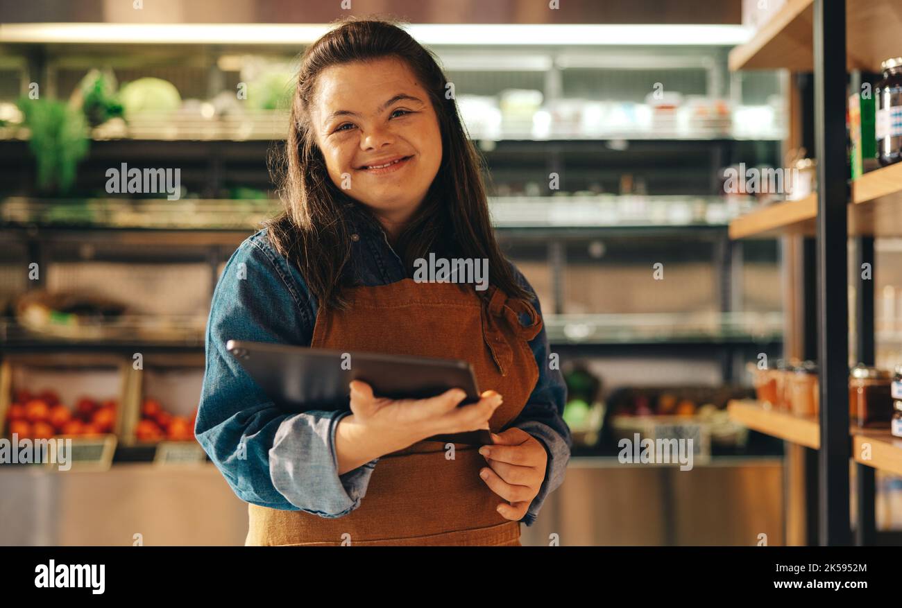 Acheter un employé avec le syndrome de Down sourire à la caméra tout en tenant une tablette numérique. Femme heureuse avec un handicap intellectuel travaillant comme un storik Banque D'Images