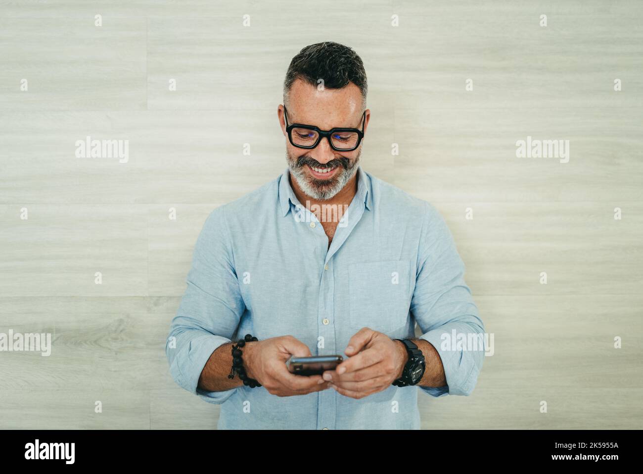 Homme d'affaires mûr souriant gaiement tout en lisant un message texte sur son smartphone. Homme d'affaires réussi à communiquer avec ses associés pendant que W Banque D'Images