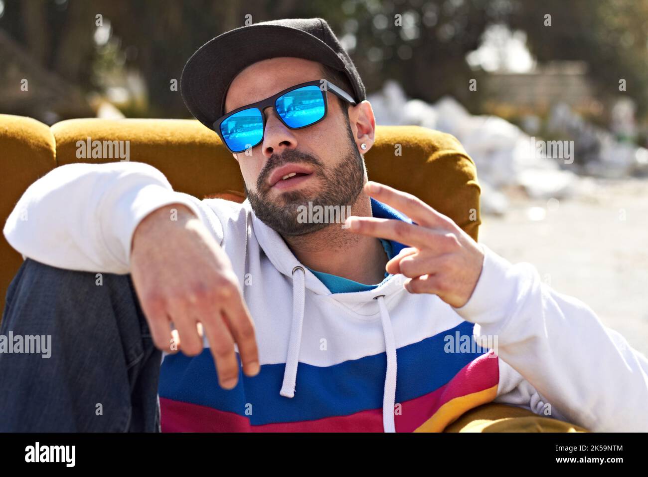La paix. Homme tendance avec des lunettes de soleil et une casquette sur un canapé dans son quartier affichant un signe de main. Banque D'Images