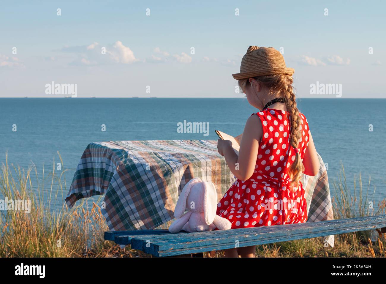 Enfant fille en chapeau et robe à pois assis sur un banc vintage lisant un livre. Enfant regardant la Sainte Bible dans les mains et priant sur la mer lanscape backgrou Banque D'Images