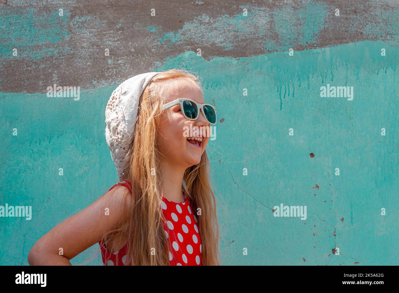 Belle fille heureuse dans une robe à pois rouges posant et souriant sur fond de mur en béton turquoise.mignon enfant joyeux avec de longs cheveux blonds chantés Banque D'Images