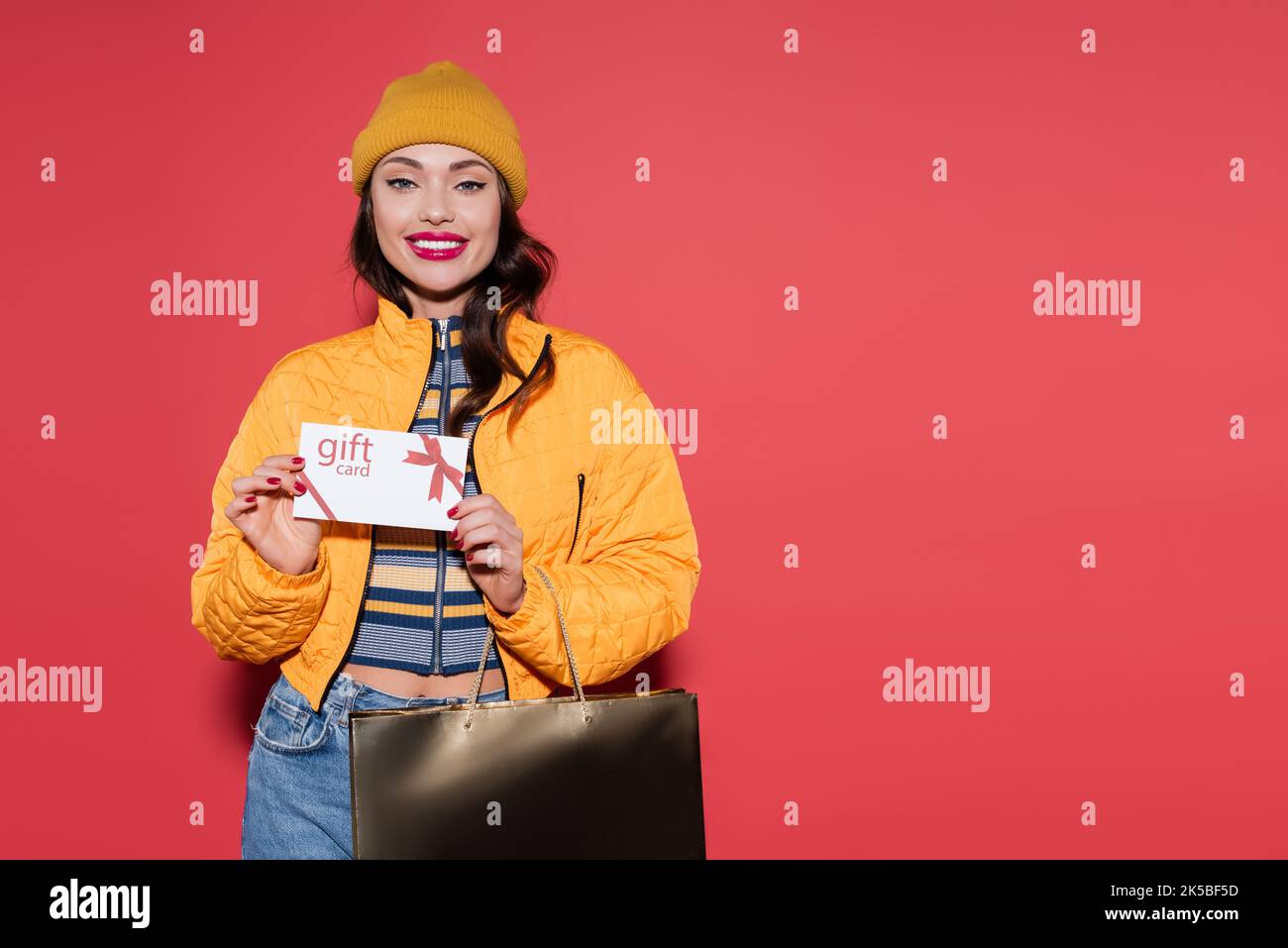 jeune femme gaie en bonnet beanie orange tenant une carte-cadeau et un sac à provisions sur rouge, image de stock Banque D'Images