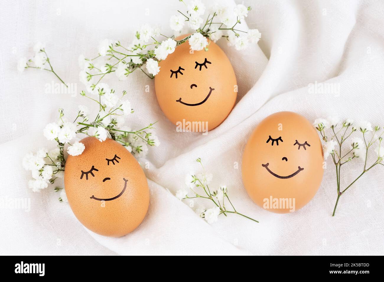 Trois œufs de Pâques heureux avec visages mignons dans des couronnes de couronne florales sur fond blanc. Œufs de Pâques avec fleurs et yeux endormis sous lumière ensoleillée. Bonne EA Banque D'Images