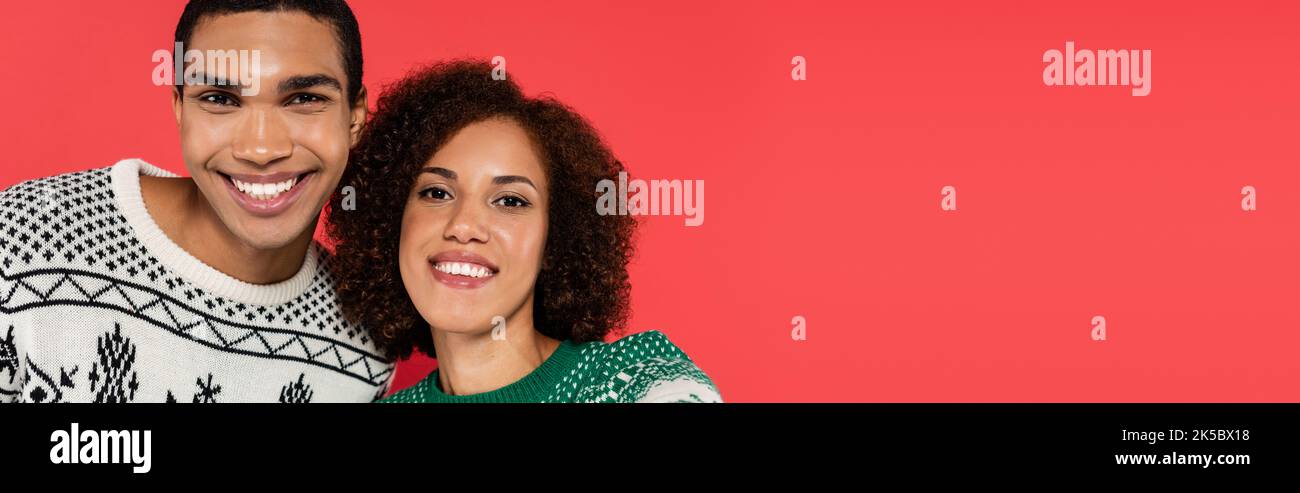 joyeux couple afro-américain en hiver pullovers souriant à la caméra isolée sur rouge, bannière, image de stock Banque D'Images