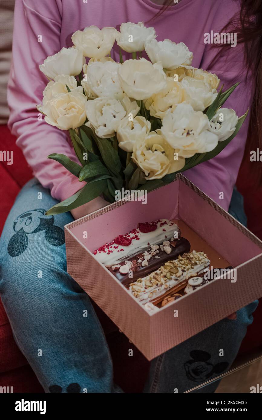 Une femme tient un bouquet blanc de fleurs et une boîte de dessert français traditionnel eclair. Femme en chandail rose et jeans avec Mickey Mouse. Banque D'Images