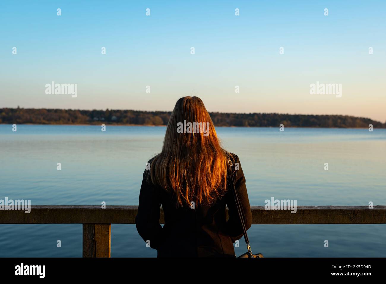 Jeune femme debout devant un lac et regardant loin. Lumière orangée au coucher du soleil qui frappe les cheveux. L'eau bleue est calme et idyllique. Banque D'Images