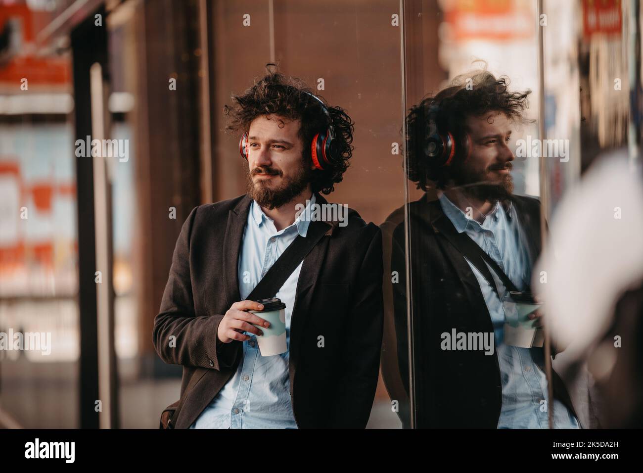 Homme d'affaires branché avec mallette en cuir dans le quartier financier à l'aide d'un smartphone tout en écoutant des chansons audio, les écouteurs boivent du café Banque D'Images