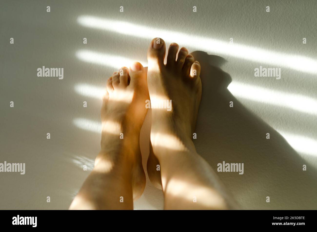 Gros plan sur les jambes d'une femme, orteils contre le mur blanc dans une maison confortable à la lumière du coucher du soleil. Ombres au mur. Bien-être Banque D'Images