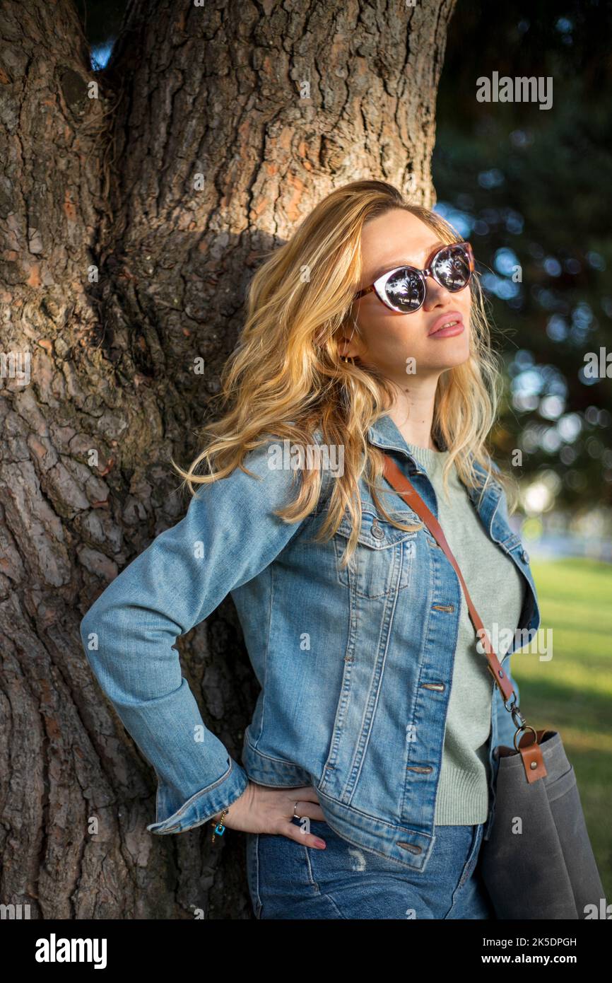 Jeune femme blonde élégante et souriante, penchée contre un arbre dans la nature Banque D'Images