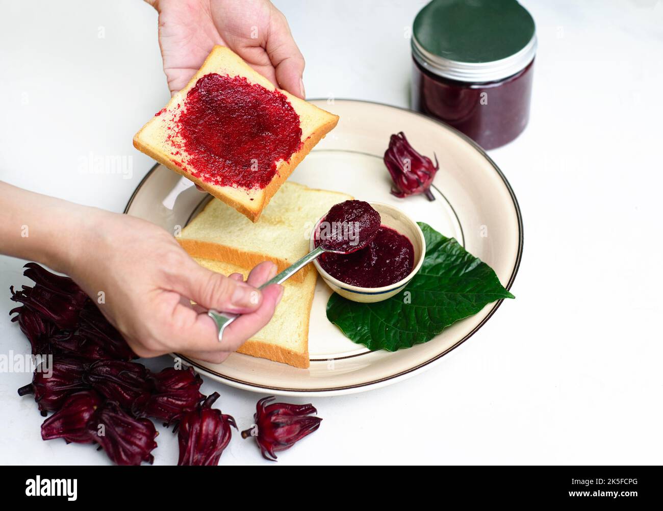 Les mains répandant la confiture d'hibiscus sur un toast contre le pot avec la confiture et les fleurs d'hibiscus Banque D'Images
