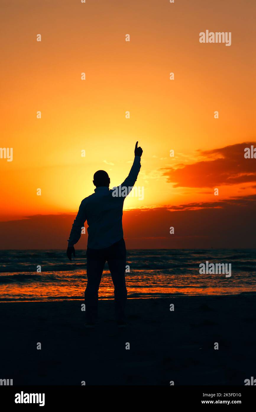 une silhouette d'un homme pointant vers le ciel pendant le coucher du soleil au bord de la mer. Banque D'Images