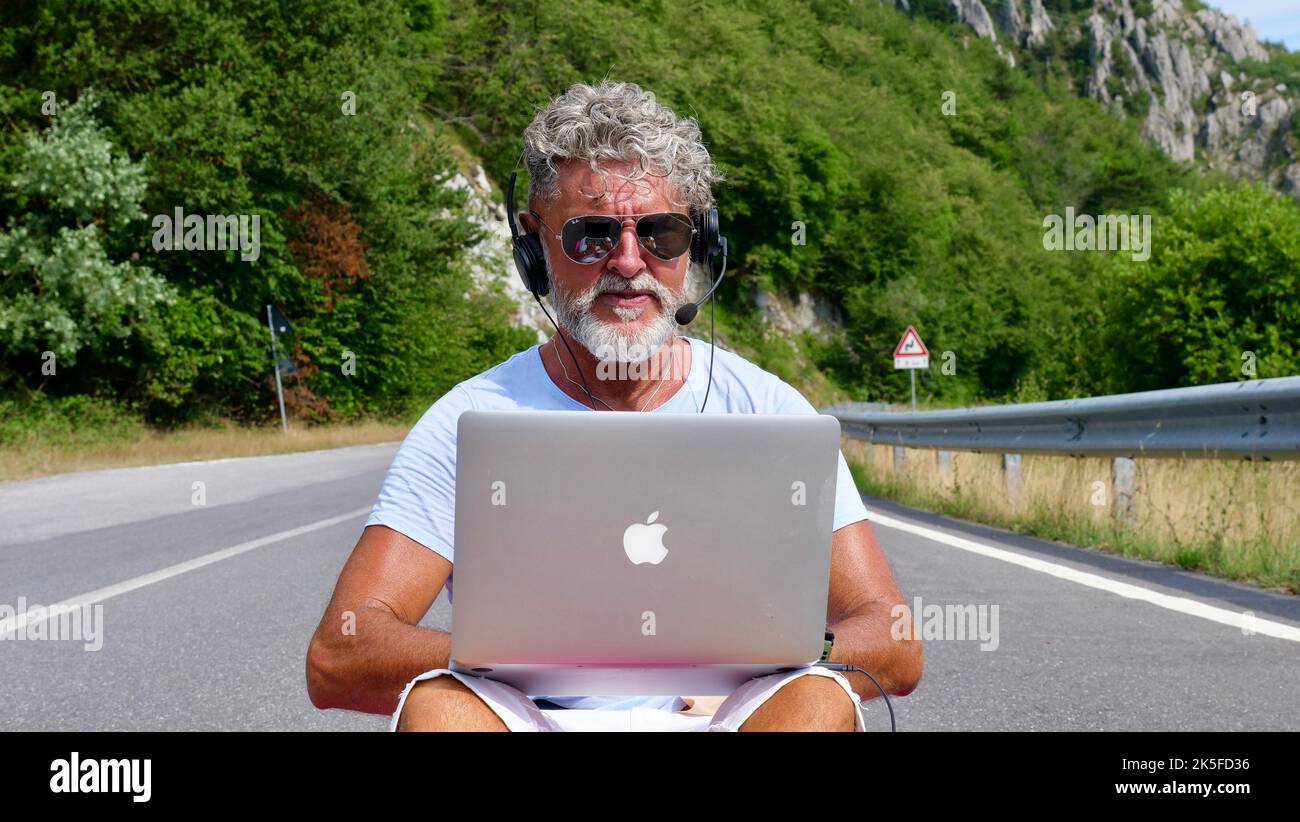 Homme âgé aux cheveux gris indépendant avec une barbe en lunettes de soleil, travaillant sur un ordinateur portable sur la nature, la route, les montagnes. Un vieil homme fou et extraordinaire est assis sur un tapis de yoga en été Banque D'Images
