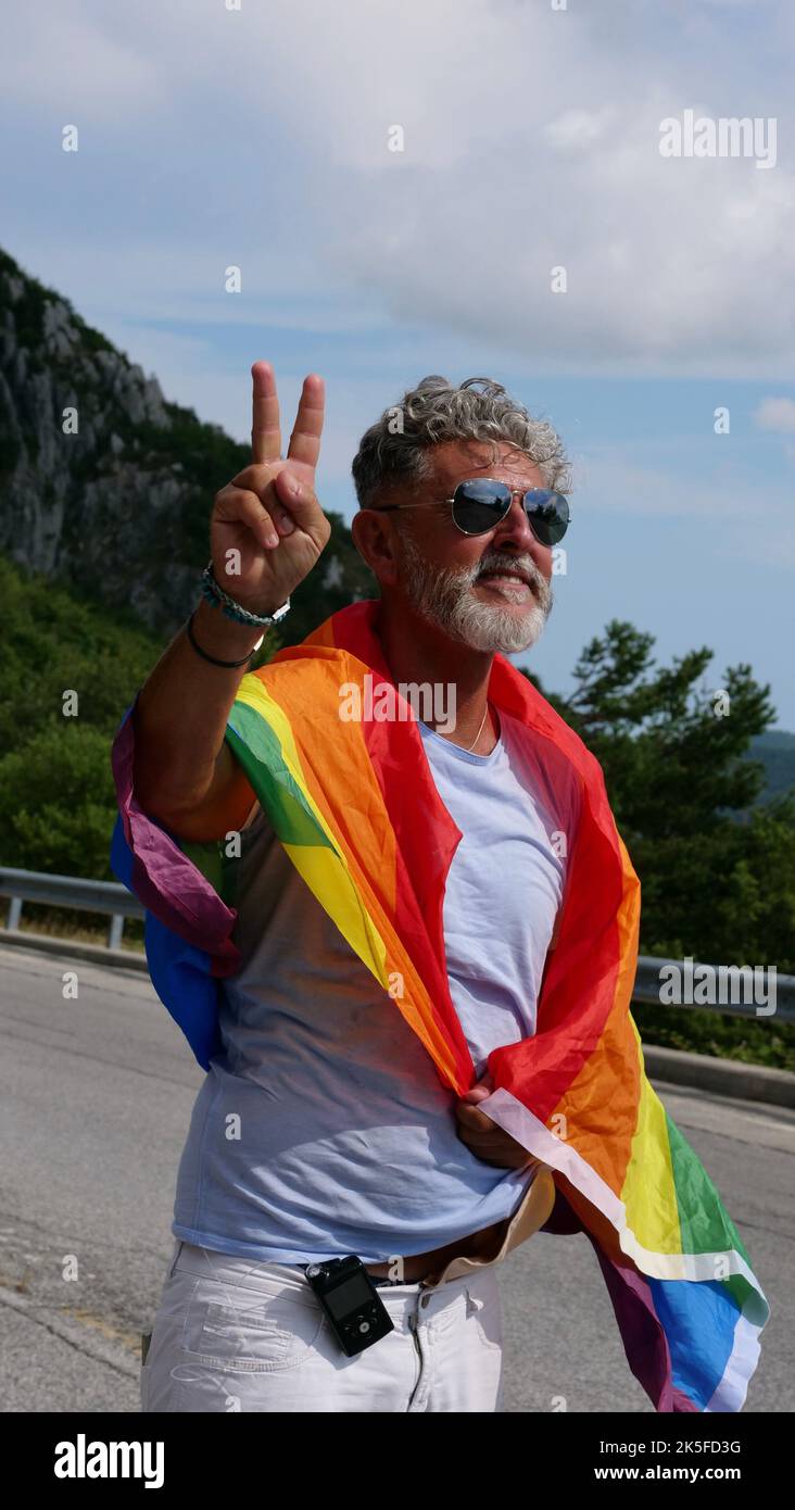 Portrait d'un homme caucasien âgé âgé âgé à cheveux gris bisexualité avec une barbe et des lunettes de soleil avec un drapeau de paix LGBTQIA arc-en-ciel dans les montagnes. Célèbre le mois de la fierté, le Rainbow Flag Day, la parade des gays Banque D'Images