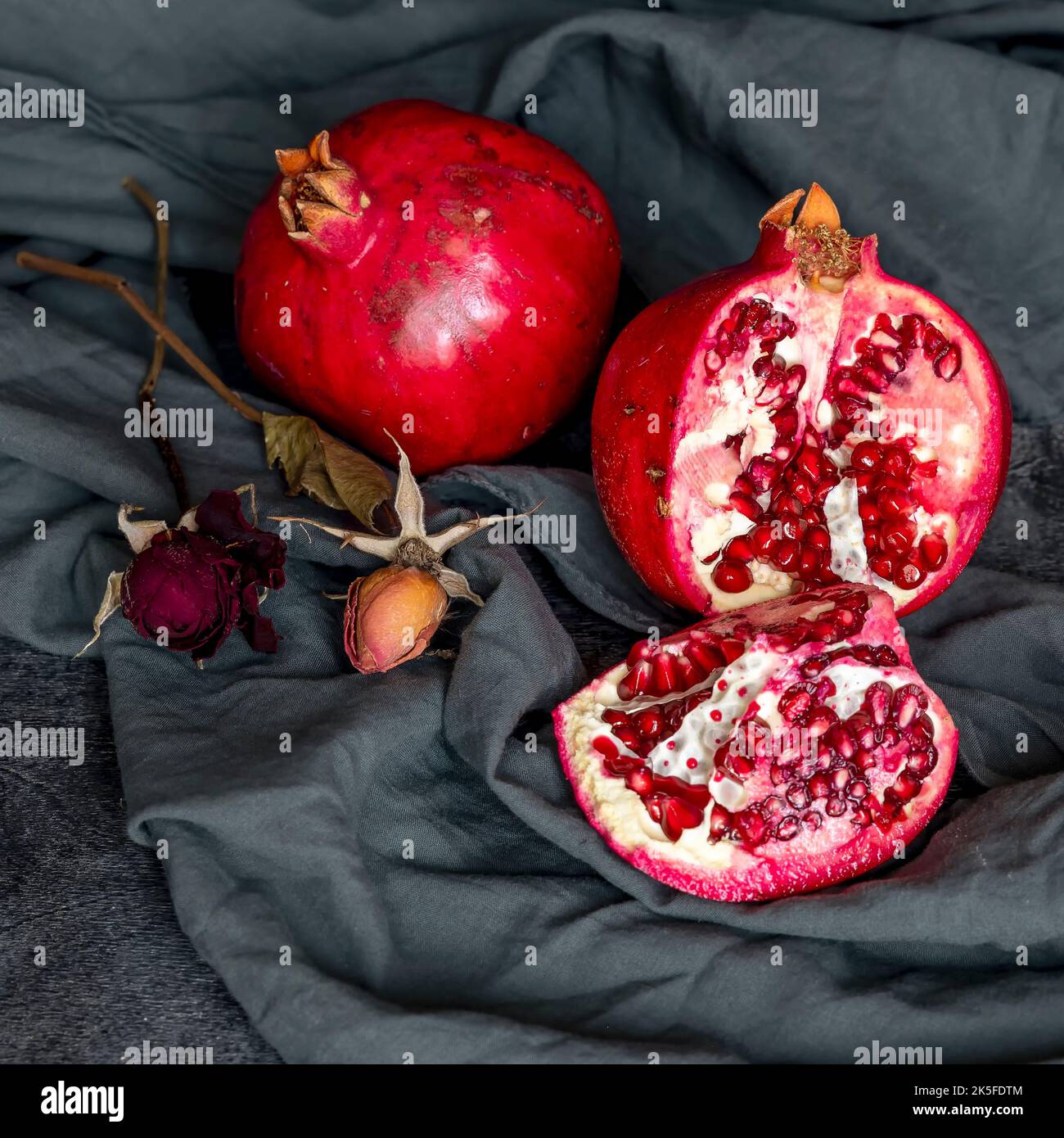 Deux grenades rouges, l'une entière et l'autre coupée en deux avec deux roses à côté, enveloppées dans un tissu gris Banque D'Images