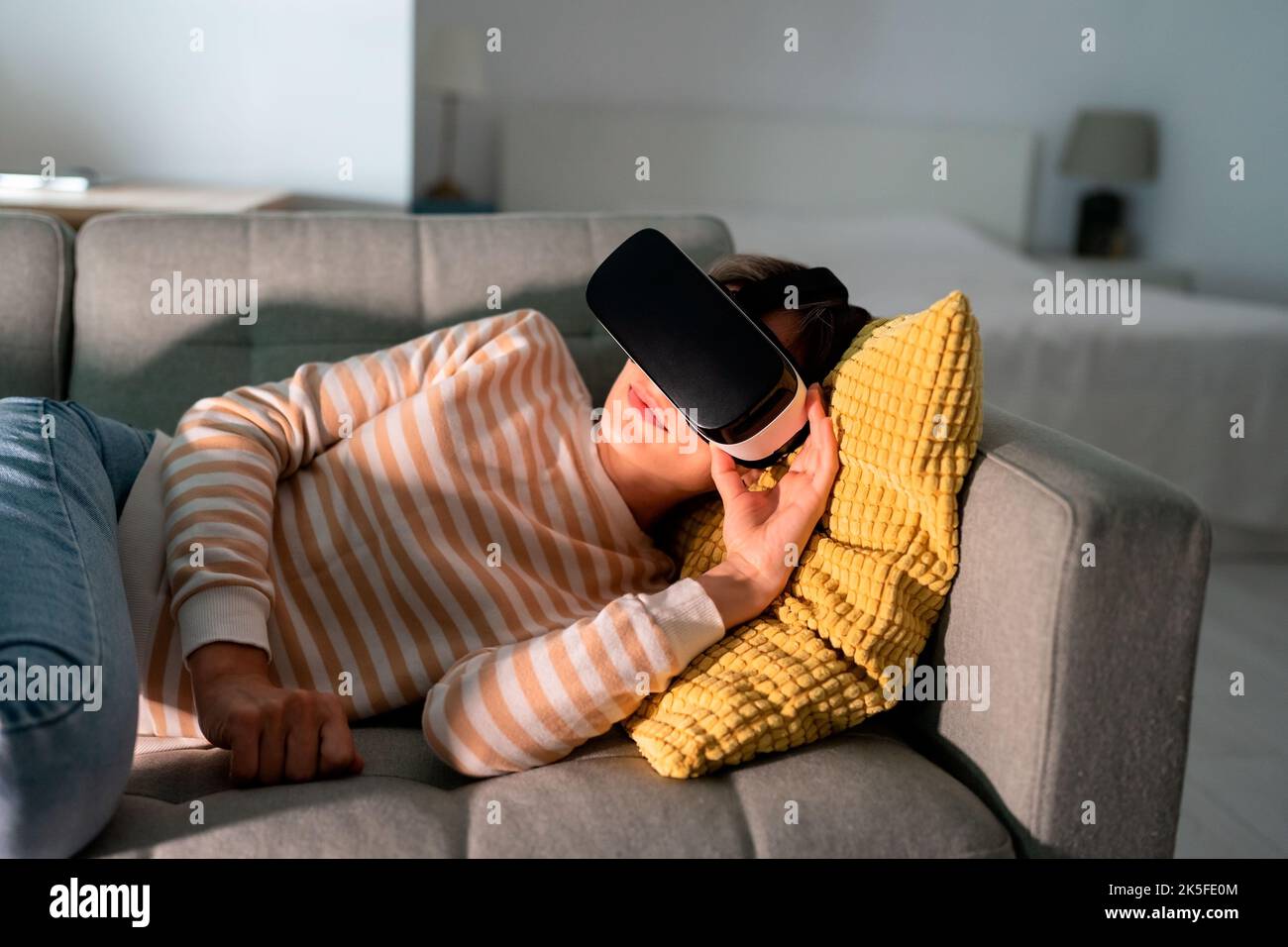 Une femme heureuse couchée sur un canapé confortable et explore un jeu intéressant dans des lunettes VR dans un appartement la nuit Banque D'Images