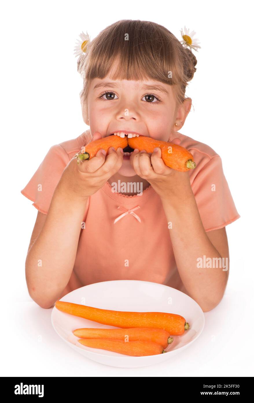Une alimentation saine. Une petite fille dans un t-shirt orange et des fleurs dans ses cheveux mange une carotte sur un fond blanc Banque D'Images