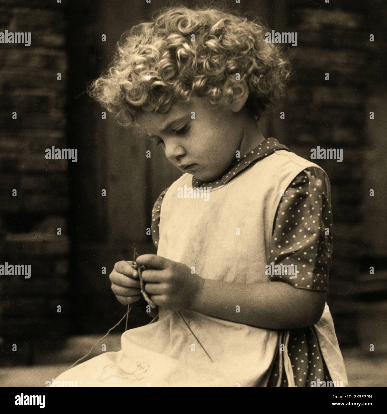 Une jeune fille avec une vadrouille en boucles porte une robe à pois et une robe chasuble comme elle est assise tricoter. Détails carrés aux tons sépia d'une photographie de portrait monochrome vintage par une photographe féminine, Kathleen Blenkhorn ARPS, de Birkby, Huddersfield, West Yorkshire. L'image originale a été prise sur film noir et blanc de format moyen à l'aide d'un appareil photo Rolleiflex TLR (reflex à double objectif) avec un objectif de prise de vue Tessar F3,5. Banque D'Images