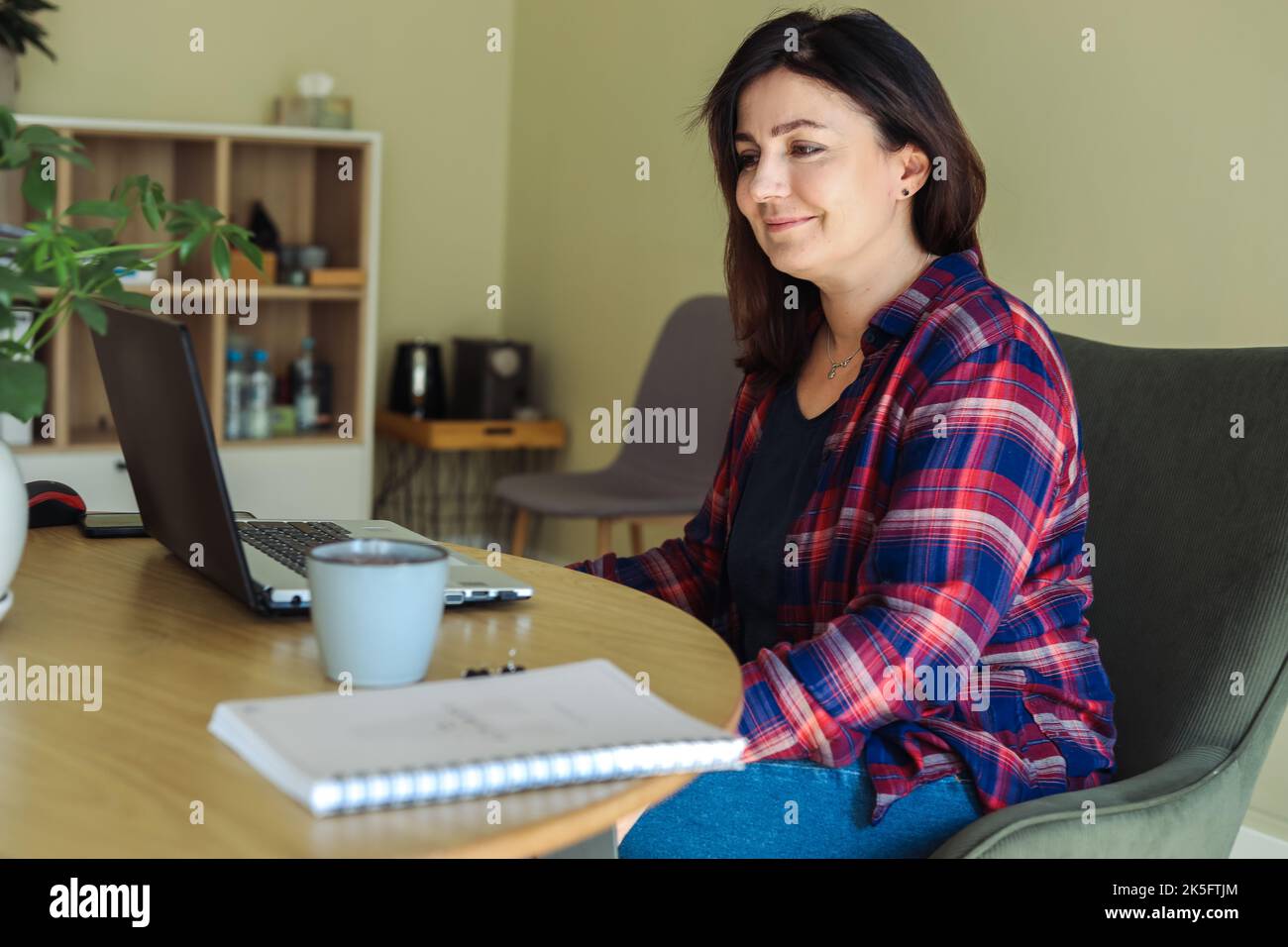 Femme souriante et confiante aux cheveux bruns dans une chemise à carreaux travail sur un ordinateur portable sur un bureau avec une tasse à café dans le bureau. Conférence du responsable Banque D'Images