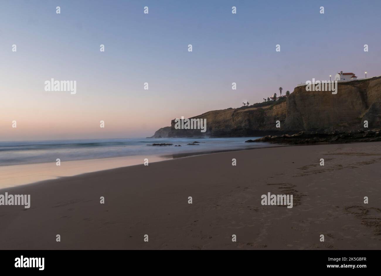 Après le coucher du soleil bleu heure de rêve longue exposition vue de plage de sable Praia da Zambujeira do Mar avec falaise avec petite chapelle et vagues de l'océan floues en rose Banque D'Images