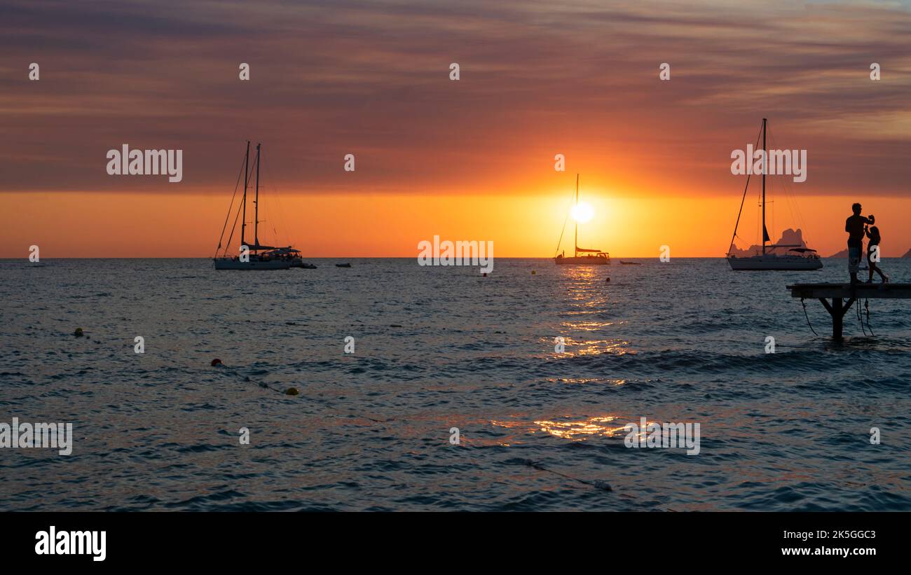 Silhouette de deux personnes regardant le coucher du soleil depuis la jetée sur la mer avec des voiliers ancrés sur l'île de Formentera en Espagne Banque D'Images