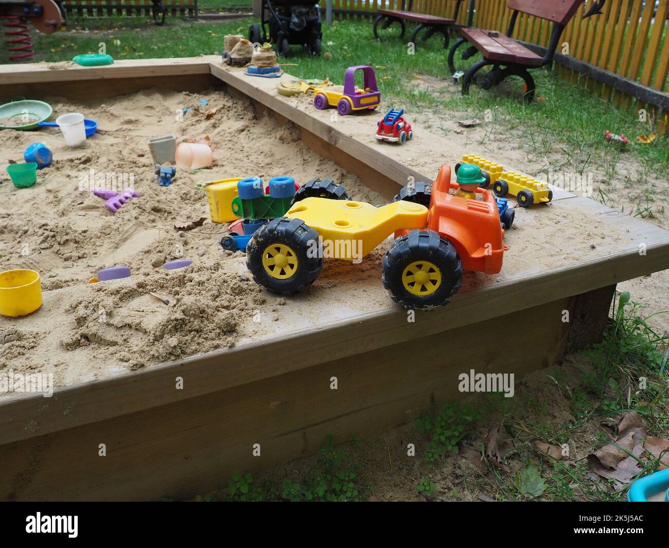 Jouet de voiture en plastique coloré sur le banc de bois d'un bac à sable sur un terrain de jeu. Autres jouets en plastique, tartes à boue, râteaux, pelles, seaux en arrière-plan. Banque D'Images