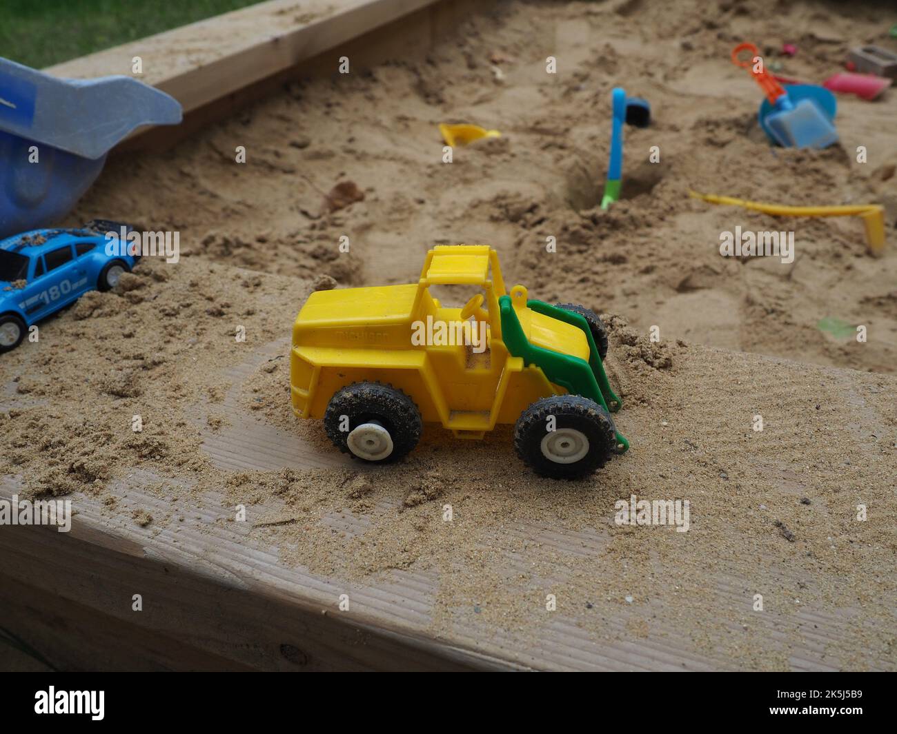 Adorable, minuscule, cassé tracteur pelle hydraulique en plastique sur le banc d'un banc de sable en bois avec différents jouets en plastique dans le sable en arrière-plan Banque D'Images