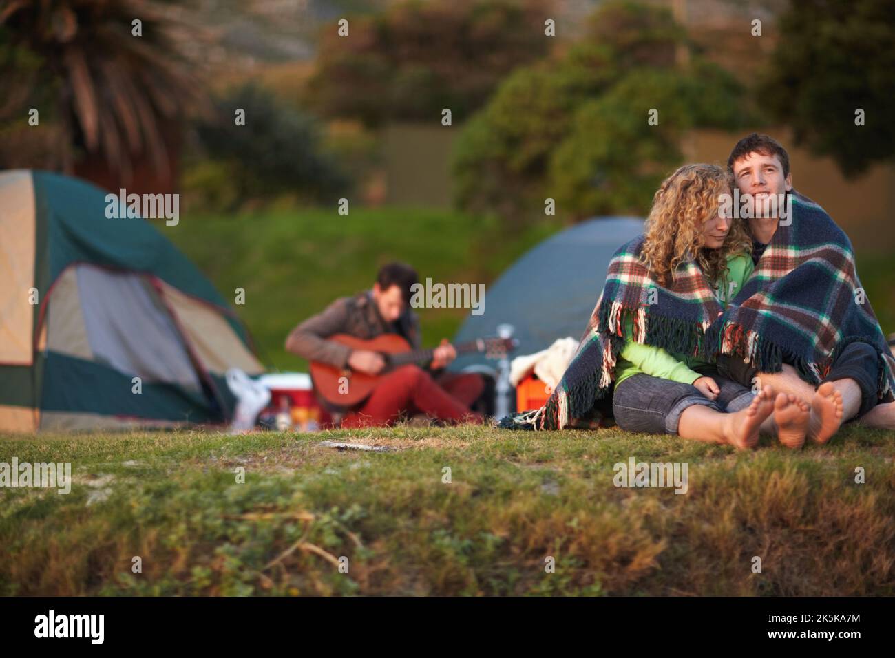 Détente sur le voyage parfait. Un jeune couple mignon enveloppé dans une couverture tout en étant assis sur une rive avec leurs amis en arrière-plan. Banque D'Images