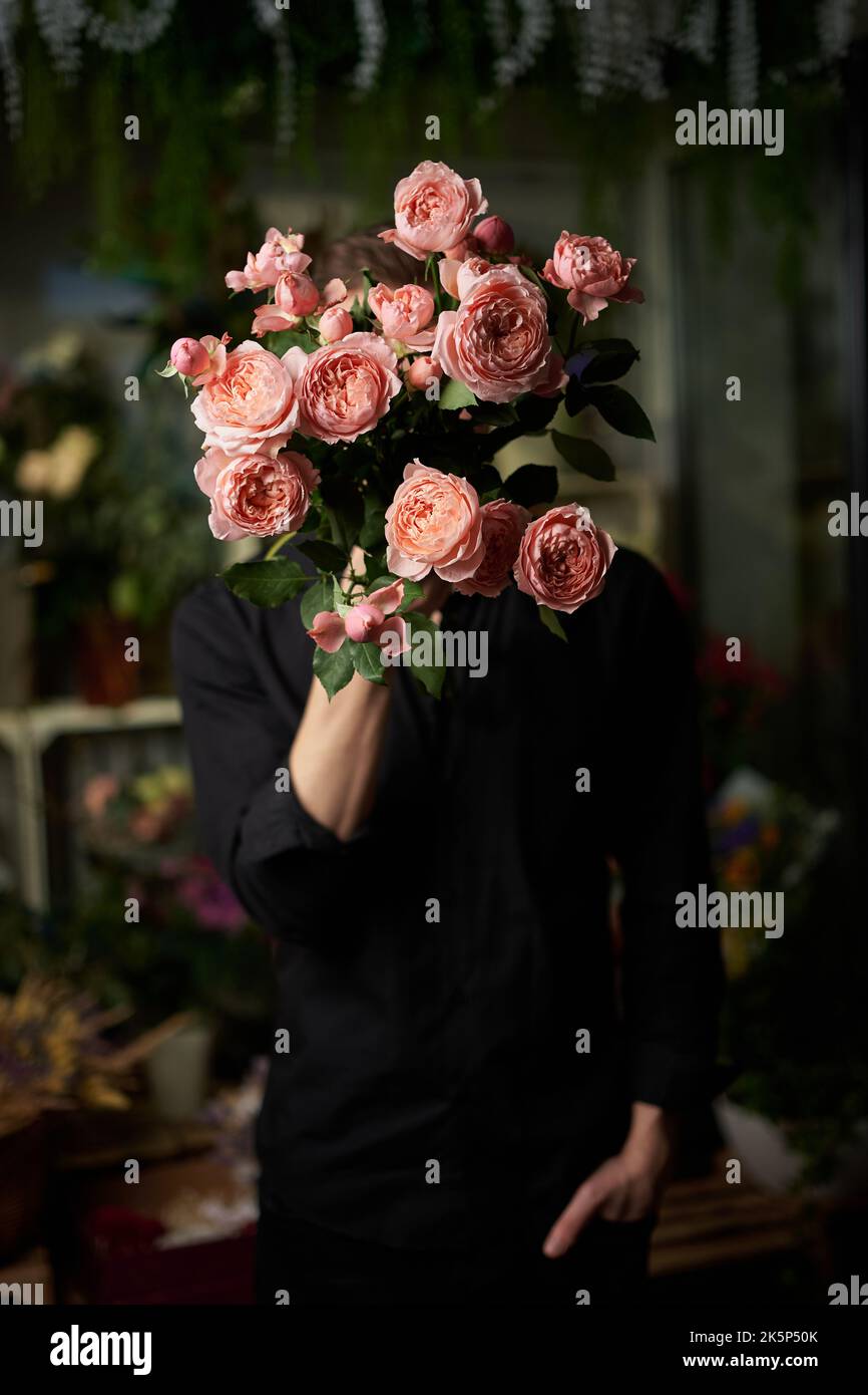 Homme en chemise noire avec bouquet de fleurs. Mignon mâle caucasien tenant un bouquet de roses fraîches devant son visage le cachant. Image clé basse. Concept amour ou romance. Photo de haute qualité Banque D'Images