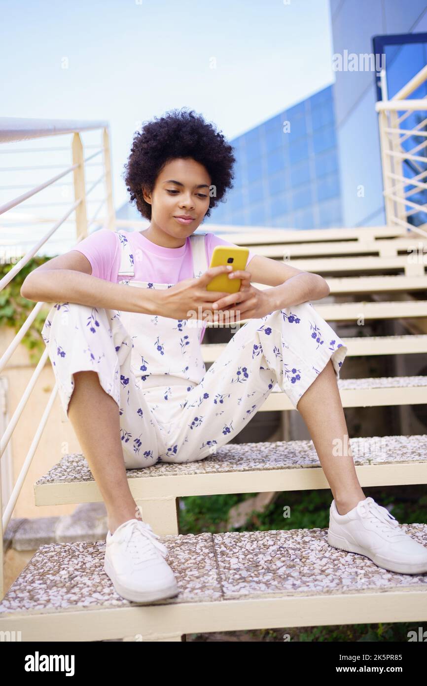 Une femme noire envoie des SMS avec son smartphone, assise sur les marches d'un bâtiment moderne. Banque D'Images