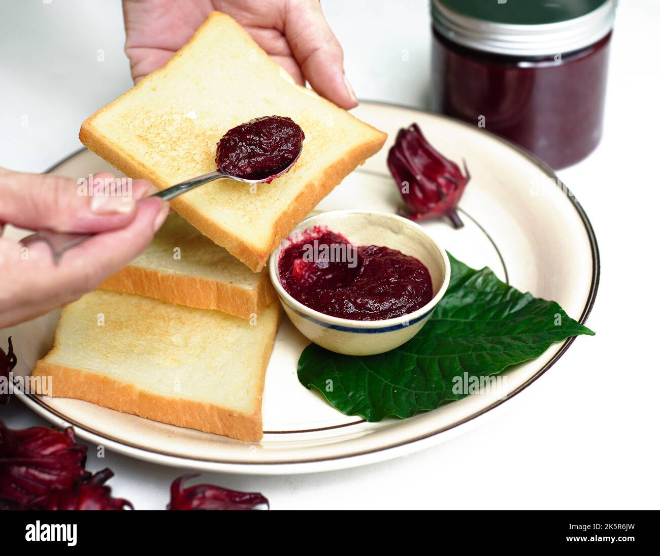 Les mains répandant la confiture d'hibiscus sur un toast contre le pot avec la confiture et les fleurs d'hibiscus Banque D'Images