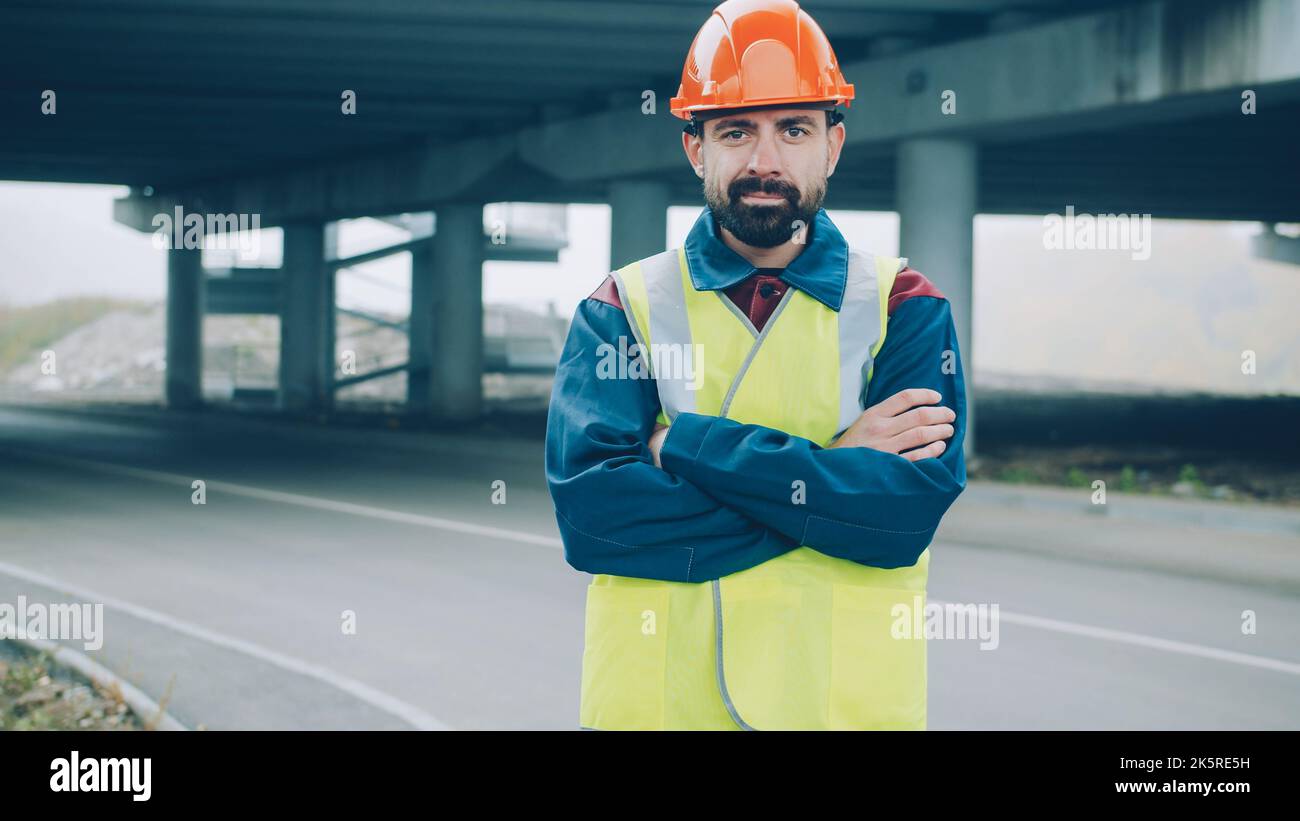 Portrait d'un jeune contremaître confiant debout à l'extérieur, portant un gilet et un casque uniformes avec un chantier de construction en arrière-plan. Concept d'entreprise de construction et de personnes. Banque D'Images
