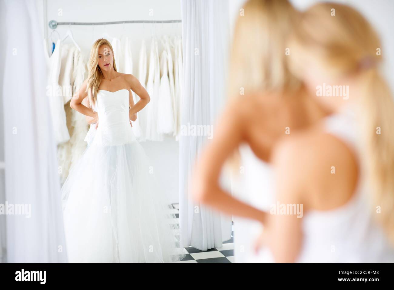 Pour un look encore plus élégant. Une jeune mariée regardant son reflet dans un miroir tout en choisissant une robe. Banque D'Images