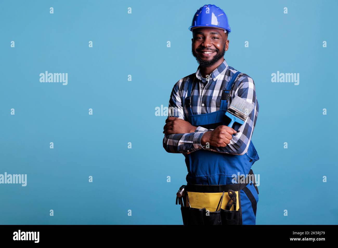 Peintre professionnel positif, homme debout avec une brosse dans les mains,  portant un uniforme de travail et un casque de protection avec les bras  croisés. Studio intérieur isolé sur fond bleu Photo