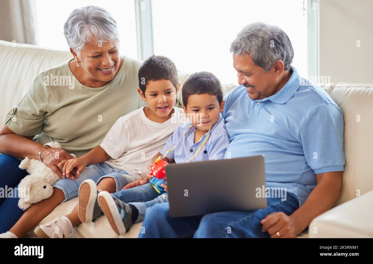 Couple senior de race mixte se liant à leurs petits-fils et utilisant un ordinateur portable sur le canapé à la maison. Homme et femme hispaniques senior s'amusant et souriant Banque D'Images