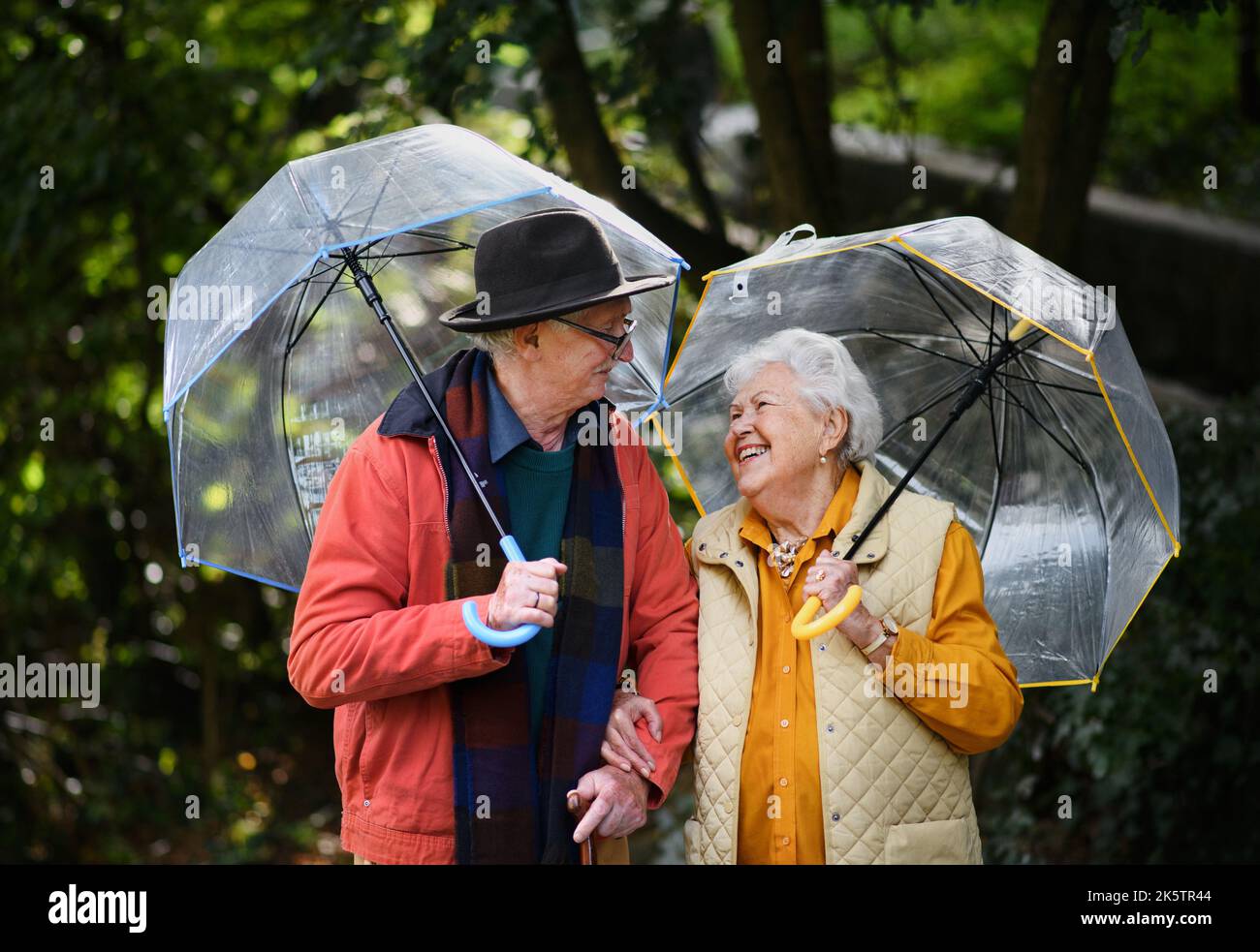 Couple de personnes âgées heureux marchant avec des parasols dans le parc de la ville ensemble. Banque D'Images