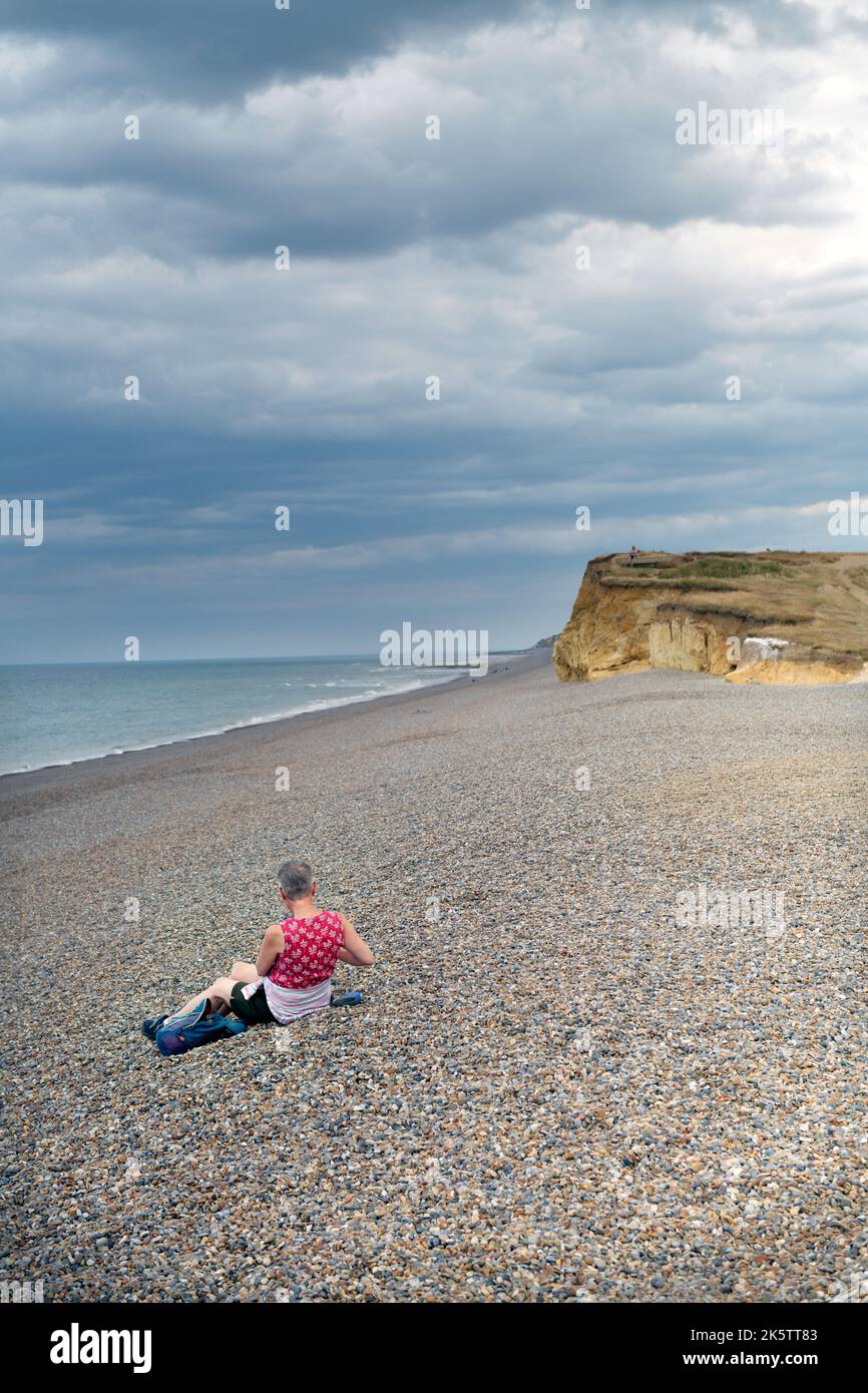 femme d'âge moyen solitaire assise sur une plage de galets à weybourne, au nord de norfolk, en angleterre Banque D'Images