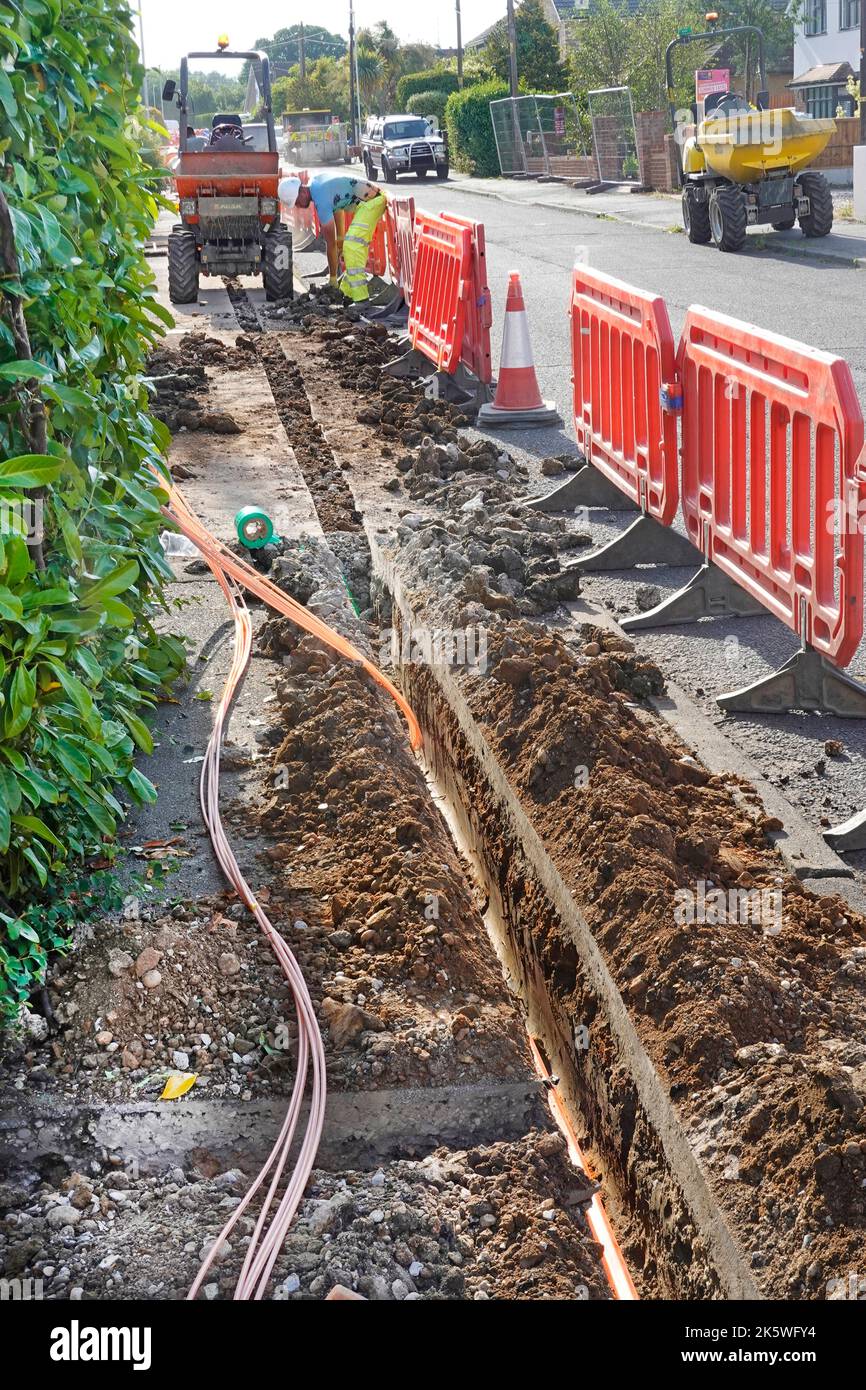 Travail en cours homme travaillant à placer de nouveaux câbles à large bande à fibre optique dans une tranchée de chaussée peu profonde derrière les barrières de sécurité des travaux routiers Essex Angleterre Royaume-Uni Banque D'Images