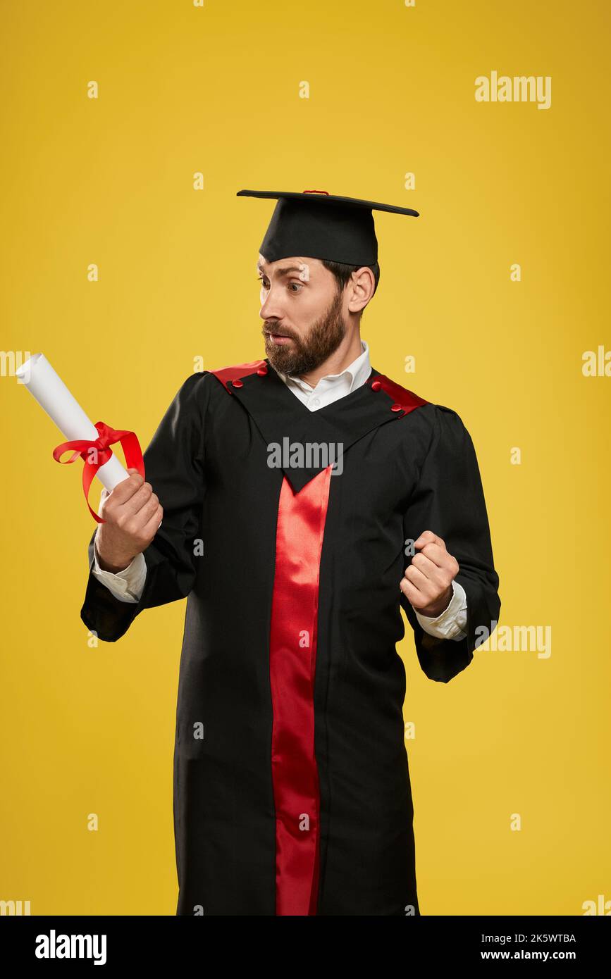 Vue de face d'un jeune homme brunette debout, regardant le diplôme. Beau garçon diplômé de l'université, université, lycée, choqué. Concept de edu Banque D'Images Vue de face d'un jeune homme brunette debout, regardant le diplôme. Beau garçon diplômé de l'université, université, lycée, choqué. Concept de edu Banque D'Images