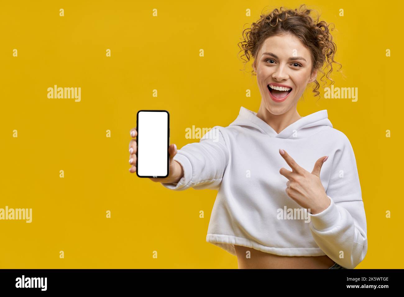Gros plan de la jolie fille souriant, tenant le smartphone. Jeune femme aux cheveux bouclés debout à la bouche ouverte, heureuse, insouciante, pointant du doigt. Concep Banque D'Images Gros plan de la jolie fille souriant, tenant le smartphone. Jeune femme aux cheveux bouclés debout à la bouche ouverte, heureuse, insouciante, pointant du doigt. Concep Banque D'Images