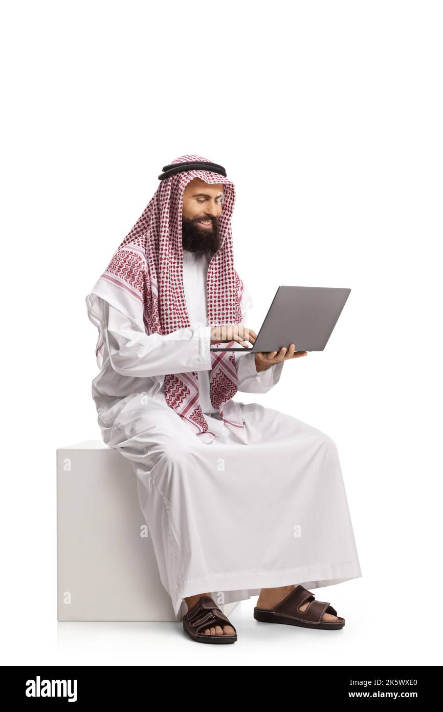 Homme arabe saoudien souriant avec un ordinateur portable installé sur un cube blanc isolé sur fond blanc Banque D'Images