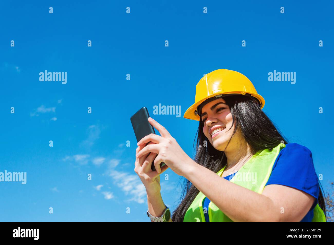 jeune latines, caucasiennes, vénézuéliennes travailleuse avec casque de sécurité jaune à l'extérieur souriant heureux de vérifier son téléphone, de lire les notifications, avec bleu Banque D'Images