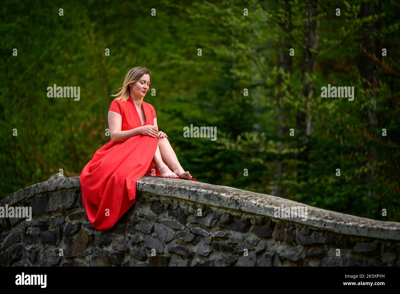 Une femme en robe rouge est assise sur un mur ancien gothique en pierre, une séance photo dans des endroits abandonnés et oubliés. Banque D'Images