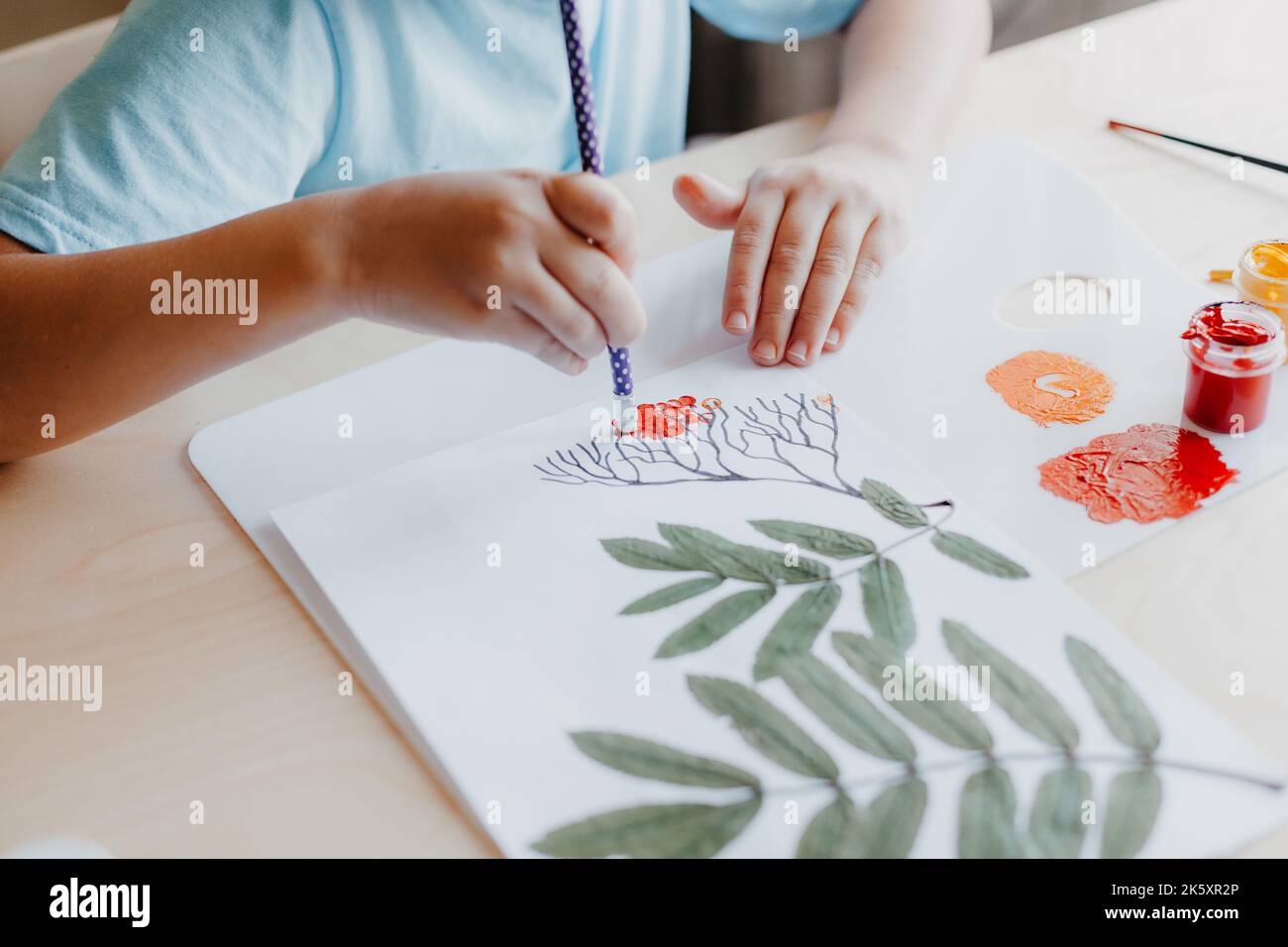 Enfant mignon assis au bureau et dessin de baies de rowanberries sur la feuille d'album avec des feuilles de rowan sèches Banque D'Images