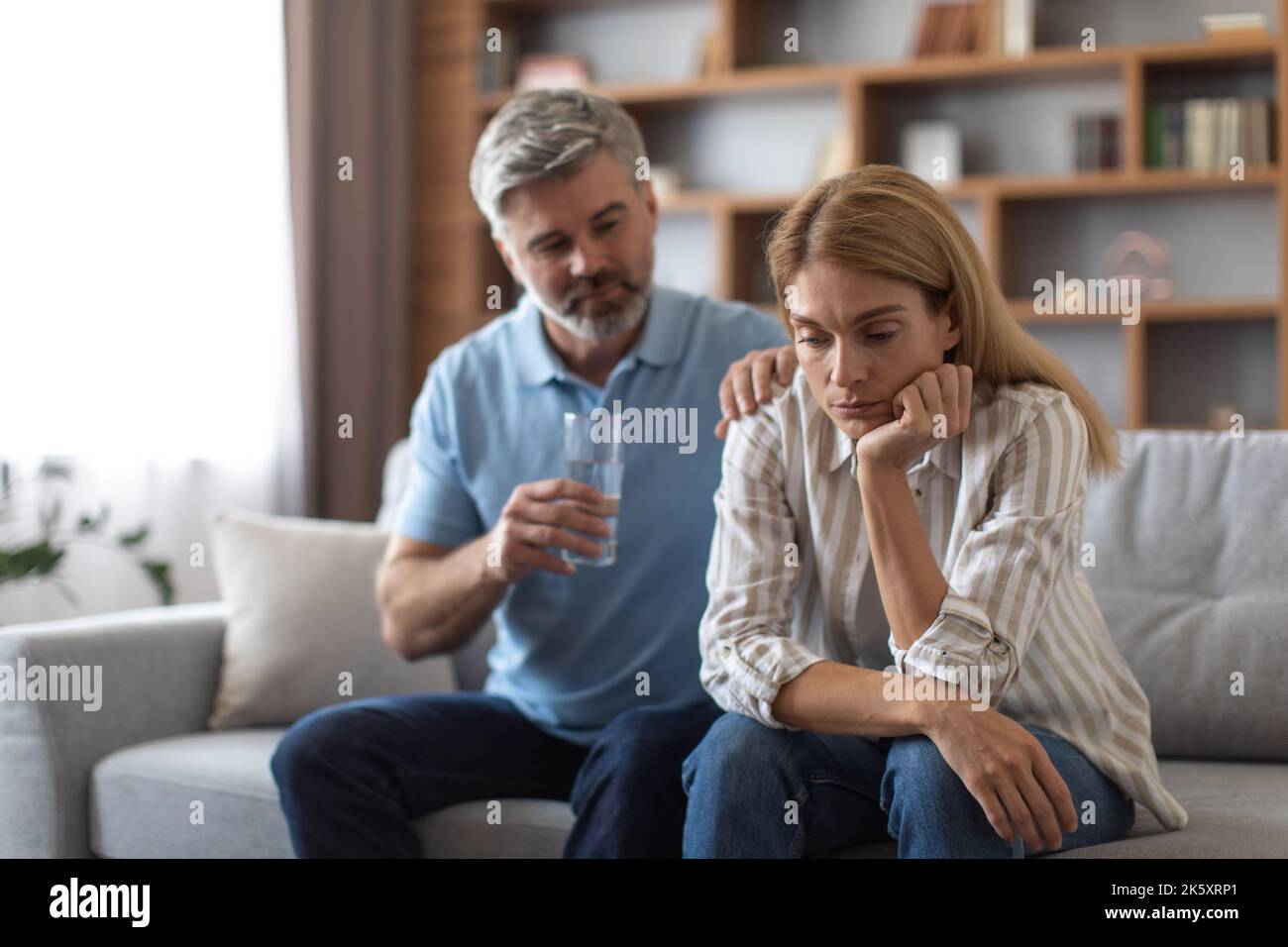Un homme européen d'âge moyen inquiétant donne un verre d'eau et calme la femme déprimée sur le canapé dans la salle de séjour Banque D'Images